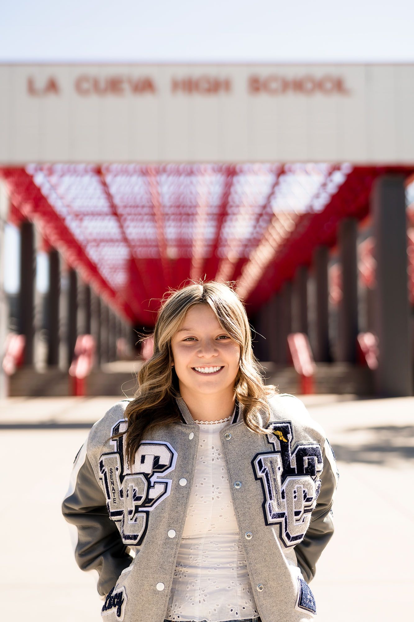 Smiling high school senior in La Cueva letterman jacket poses in front of La Cueva High School entrance.