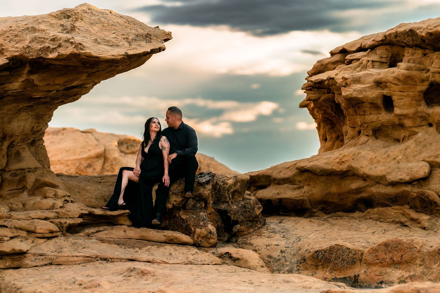 Couple sitting together between sandstone rock formations under dramatic cloudy sky.