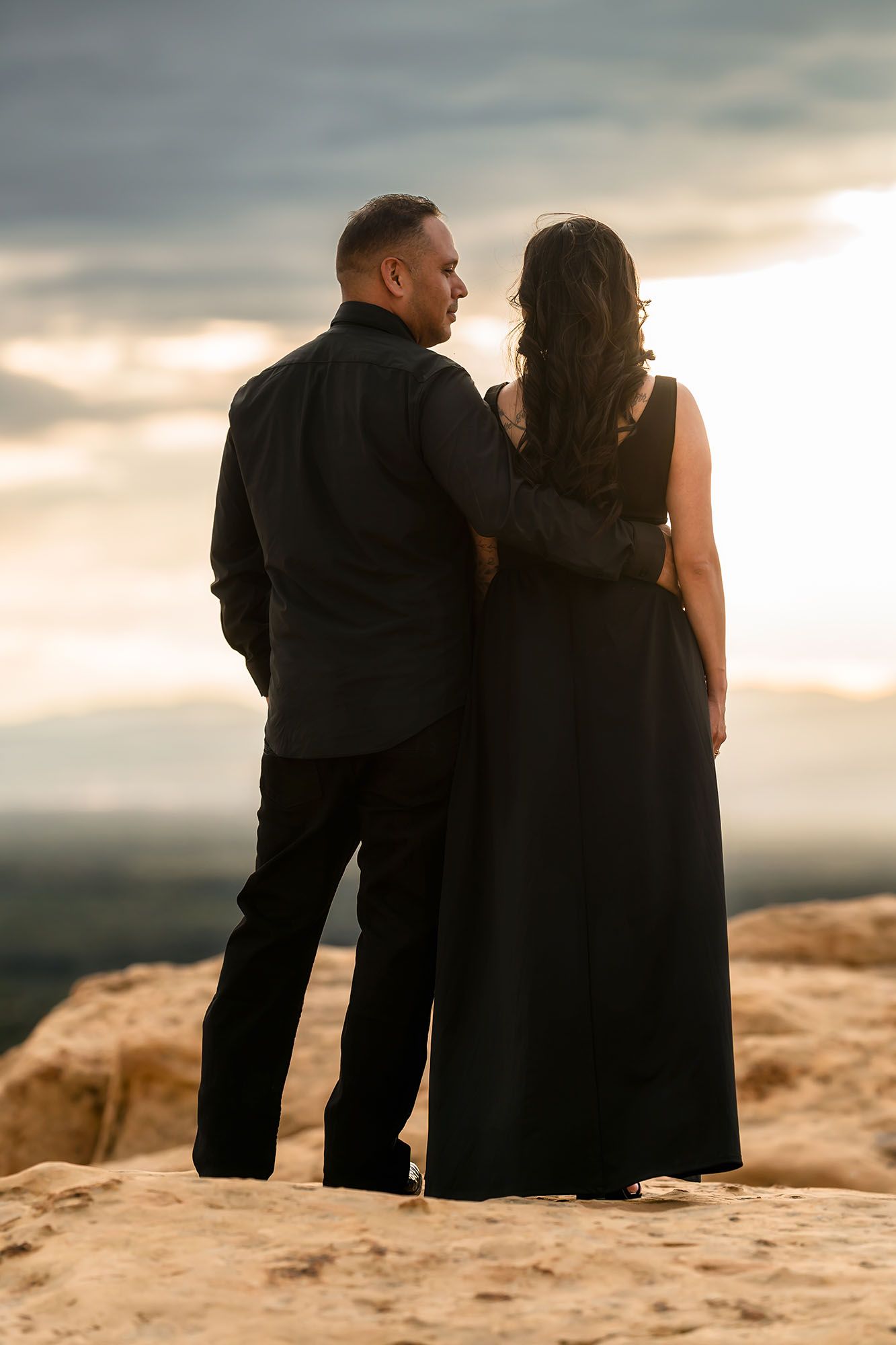 Couple in black standing arm in arm on rocky overlook gazing at a glowing sunset over vast landscape.