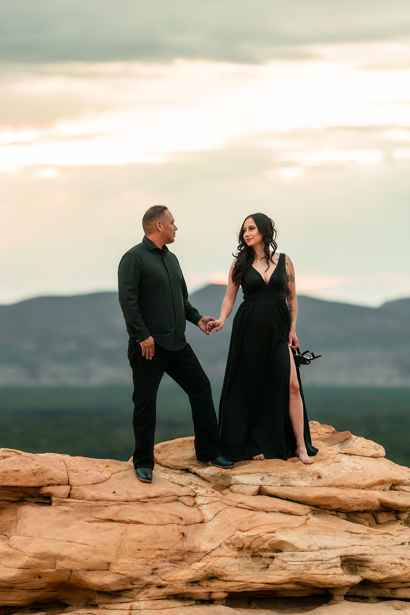 Man in black suit and pregnant woman in black gown hold hands on desert rock at dusk.