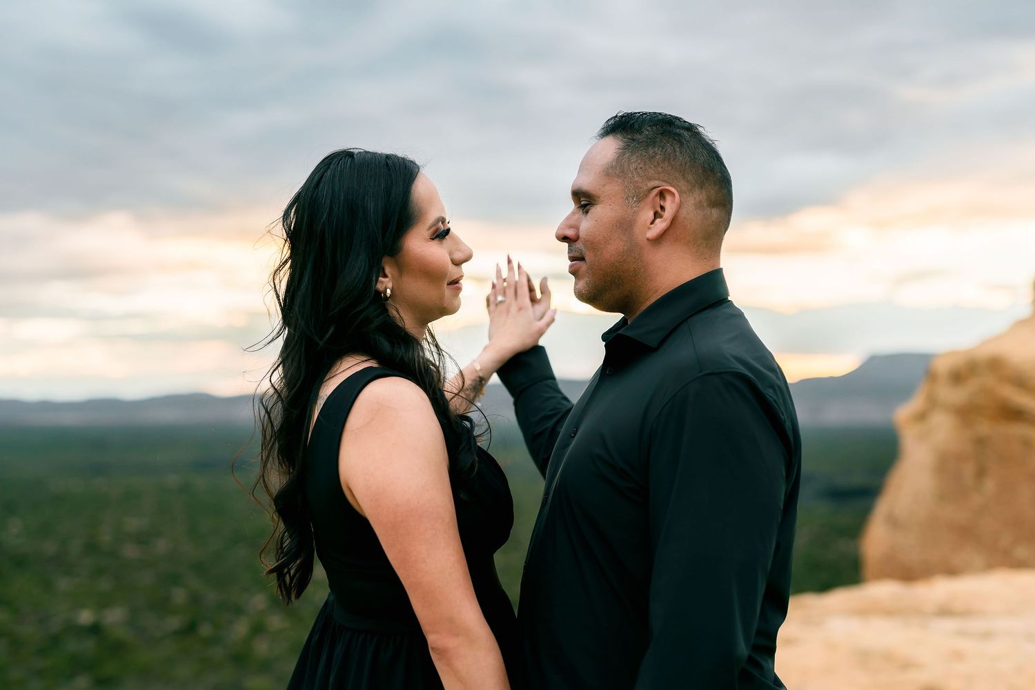 An elegant couple in black stands on a rocky overlook at dusk, gazing at each other with a ring visible.