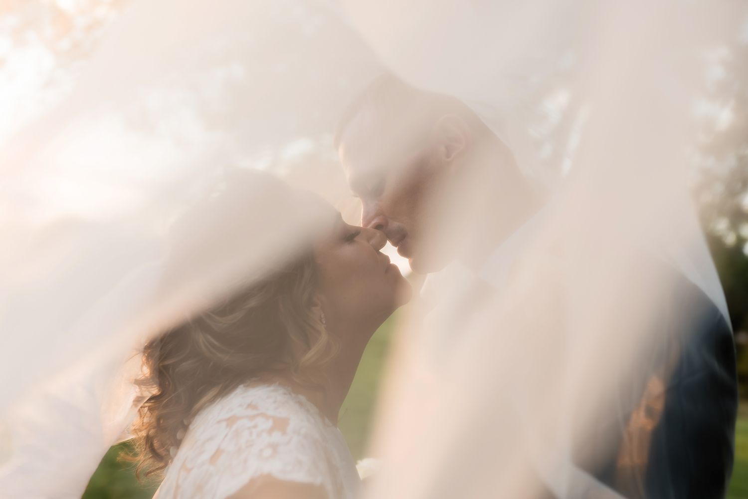 A bride and groom share a romantic kiss beneath a flowing white veil at their outdoor wedding ceremony.
