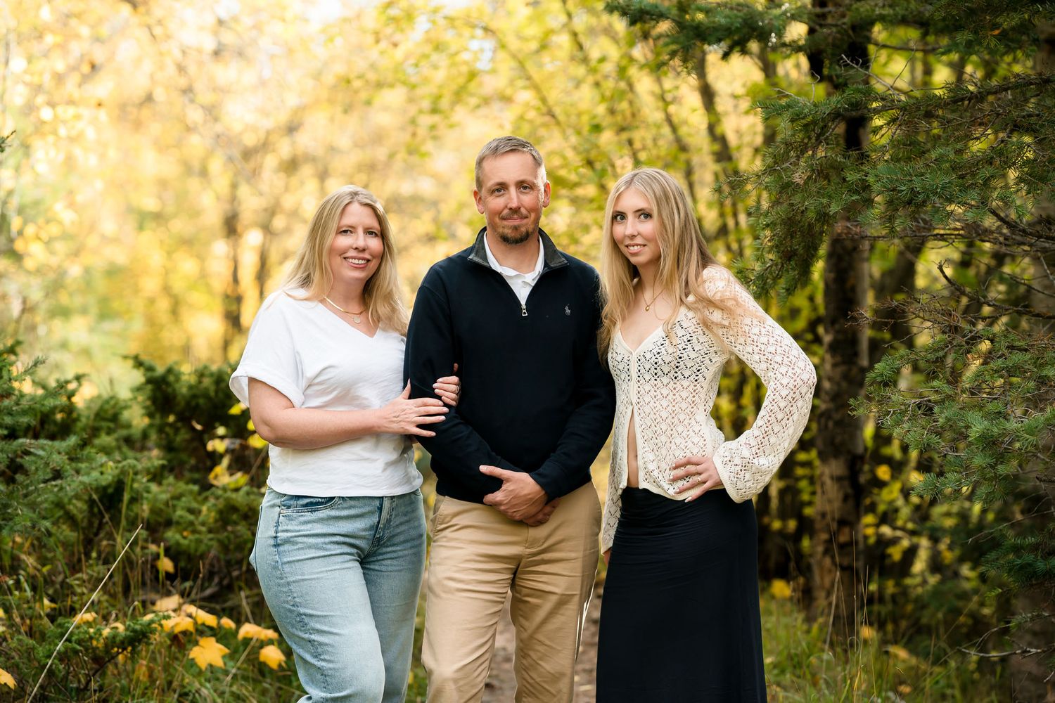Three people posing together outdoors surrounded by vibrant yellow and green autumn trees.
