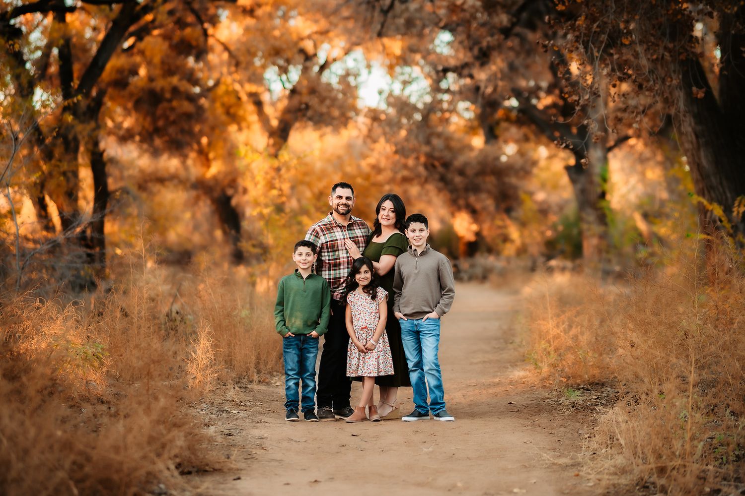 Family of six poses on a dirt path surrounded by vibrant autumn trees with golden and orange foliage.