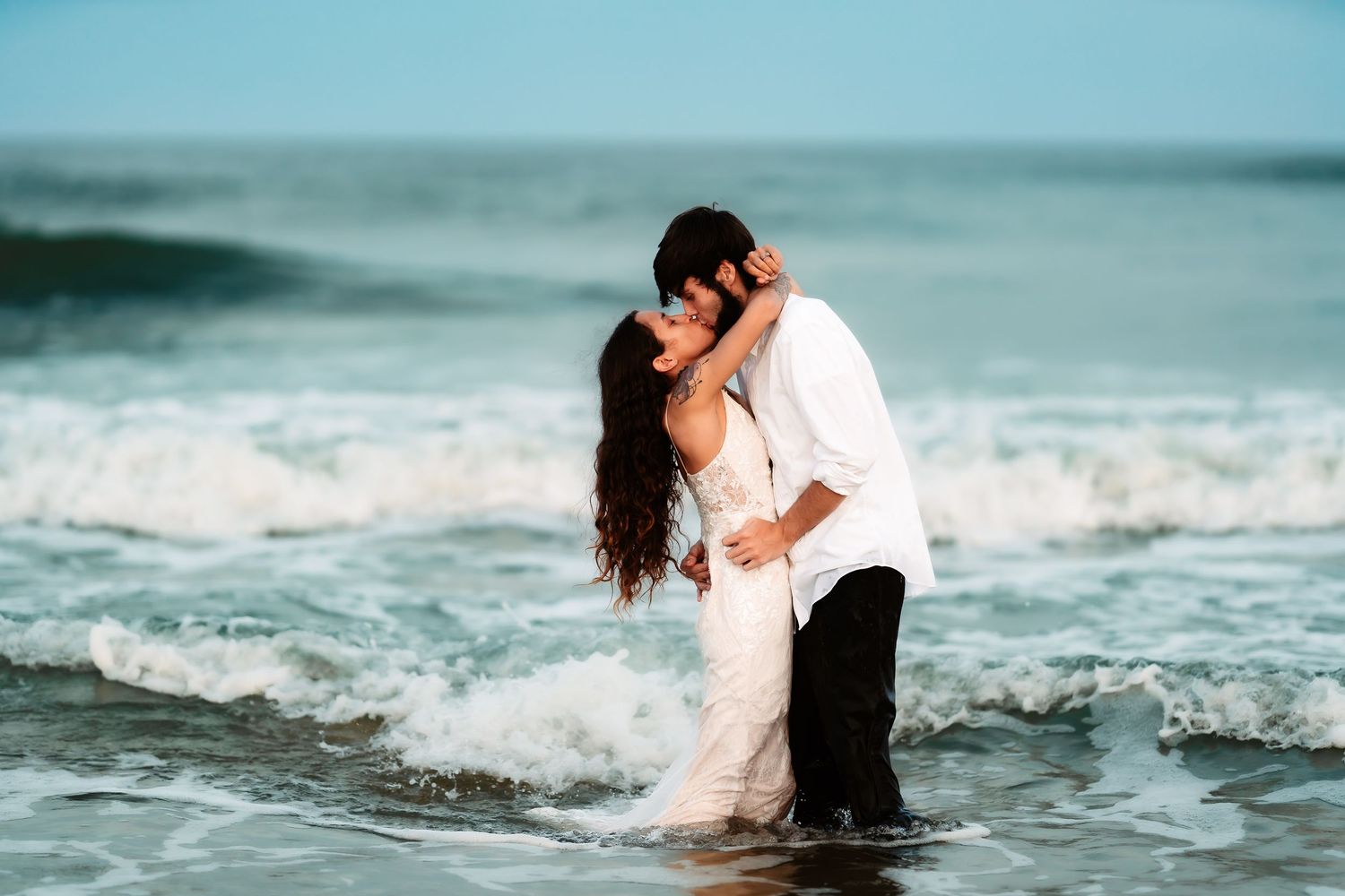 Couple embracing in ocean waves during romantic beach photoshoot with teal water background.