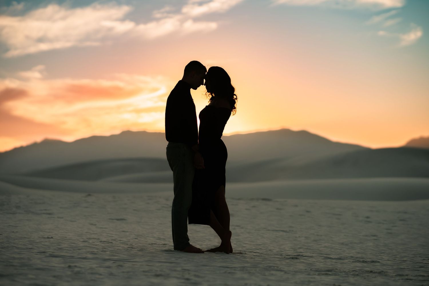 Silhouetted couple embracing in romantic pose against sunset sky over desert mountains and sand dunes.