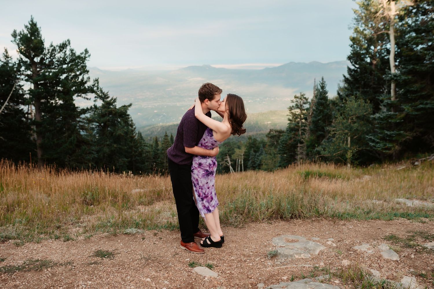 Couple sharing tender moment on mountain trail with sweeping valley views and evergreen forest.