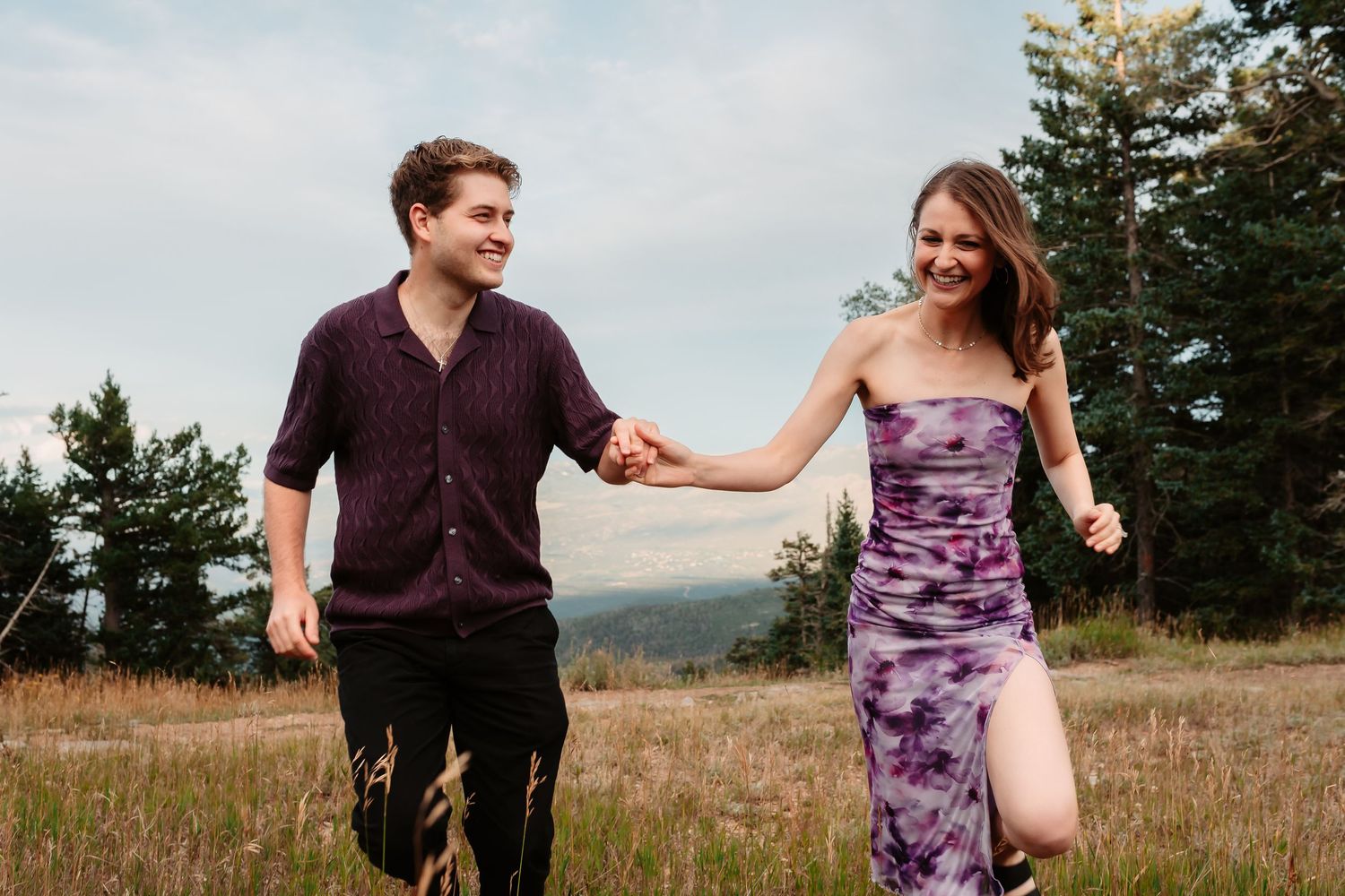 A romantic outdoor photoshoot in a mountainous field with a couple in purple and black attire dancing together.