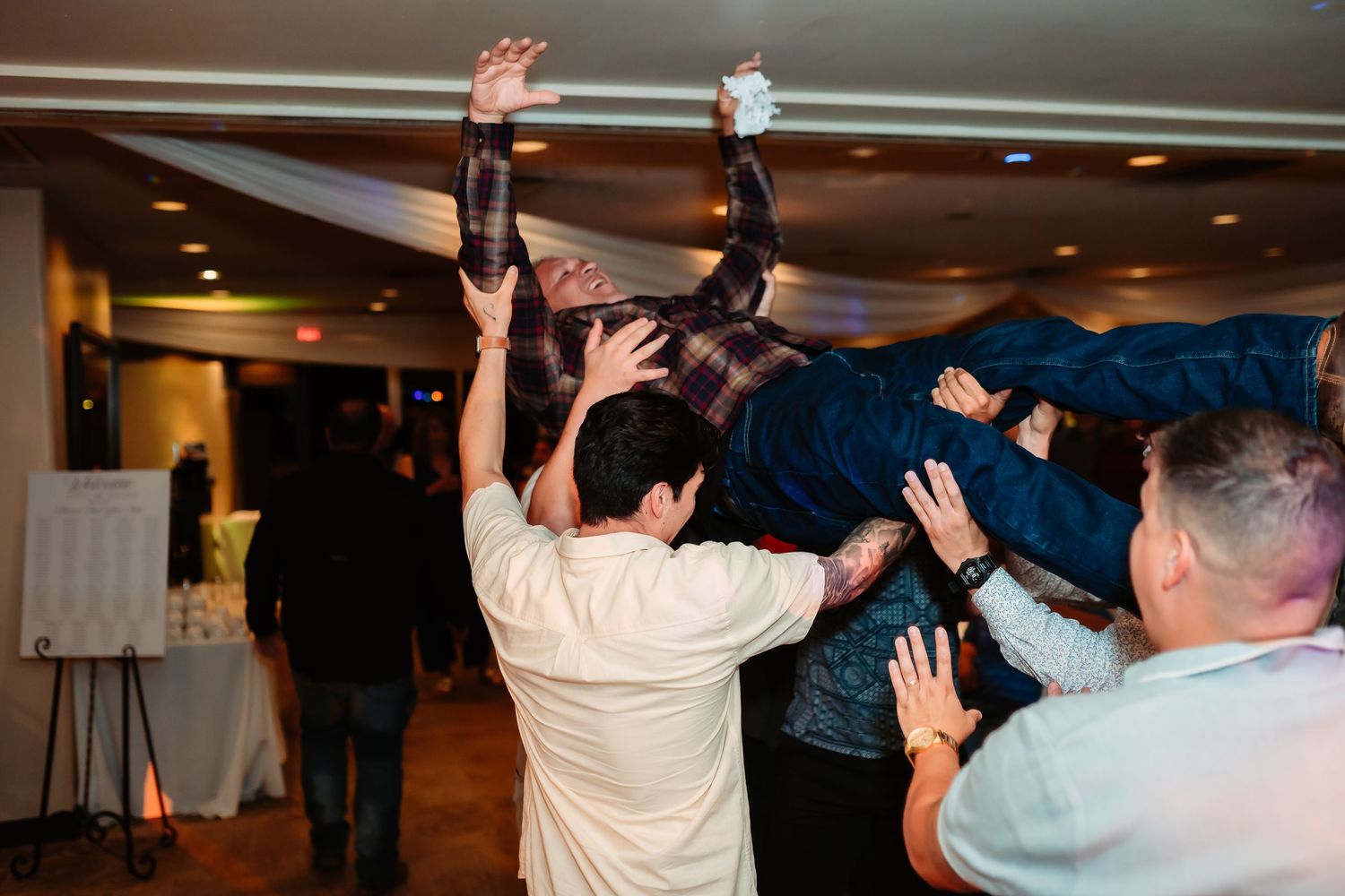 Wedding reception guests dancing and celebrating with their hands in the air at an indoor venue.