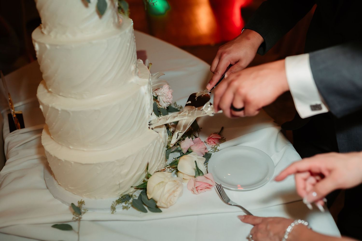 Detailed view of a pristine white wedding cake being cut with colorful lighting in the background.