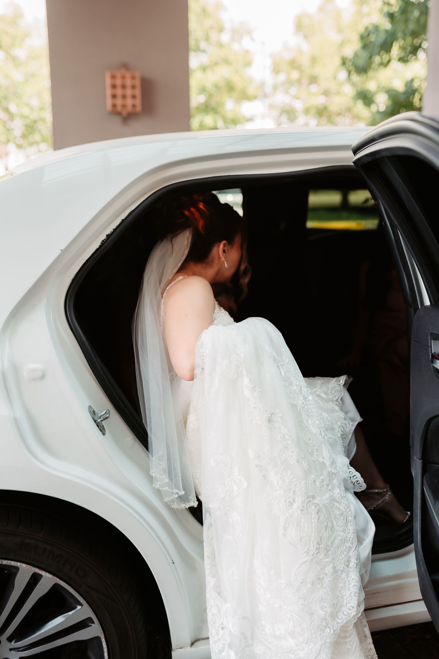 A bride in an elegant white lace gown enters a classic white wedding limousine.