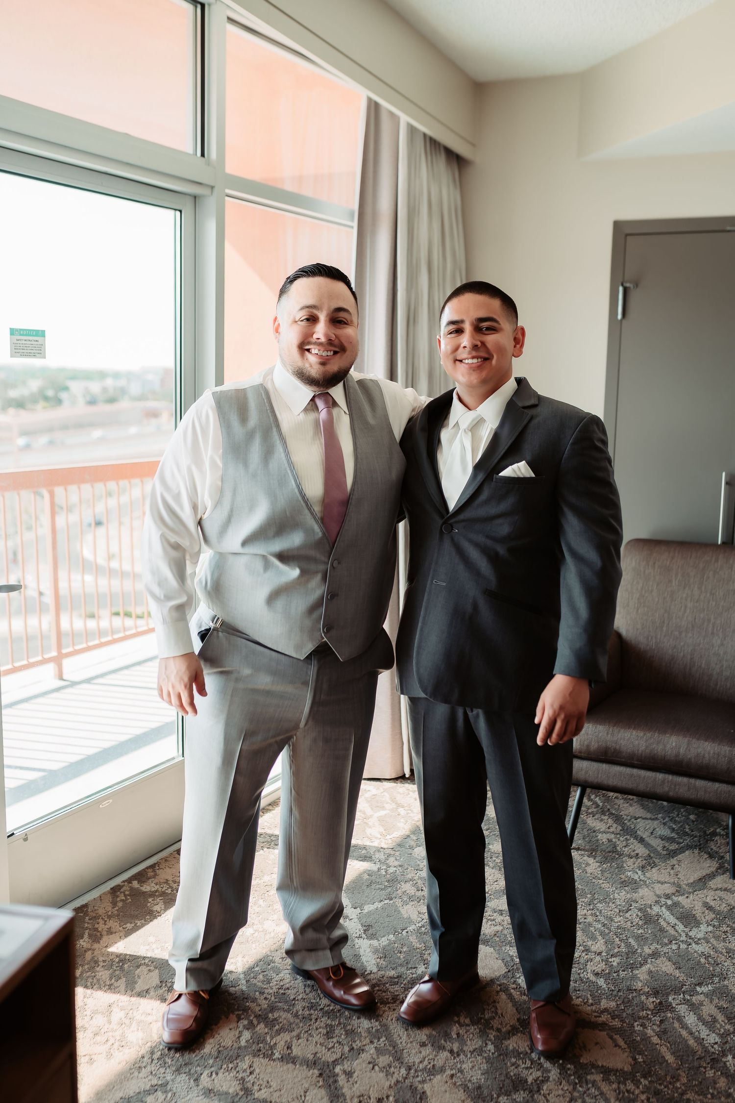 Two people in formal attire share candid moments near bright windows in a modern interior space.
