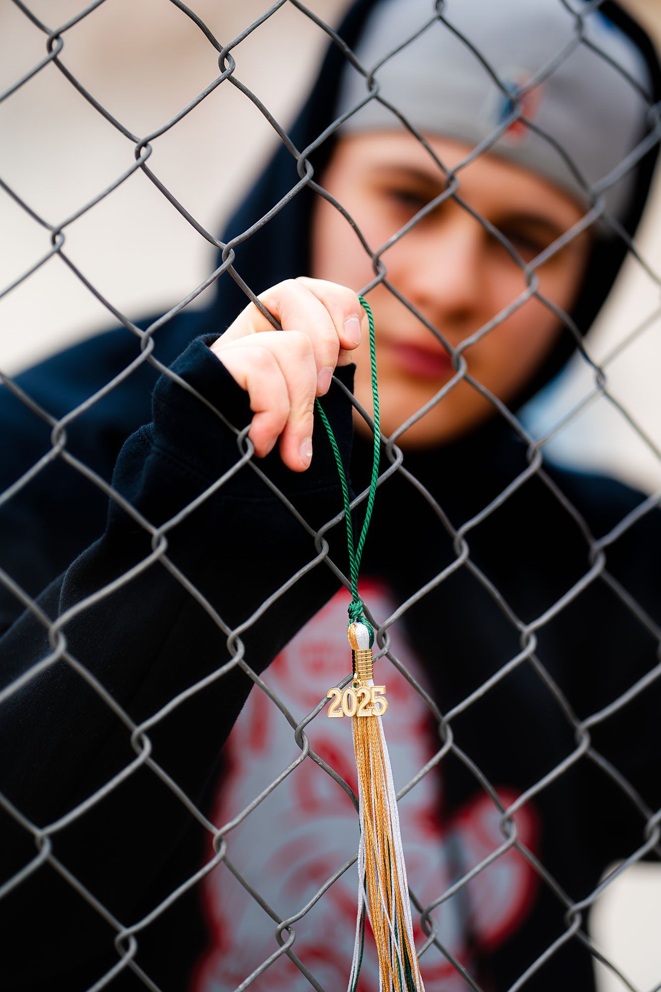 A student holds a 2025 graduation tassel on a chain-link fence, wearing a dark hoodie.