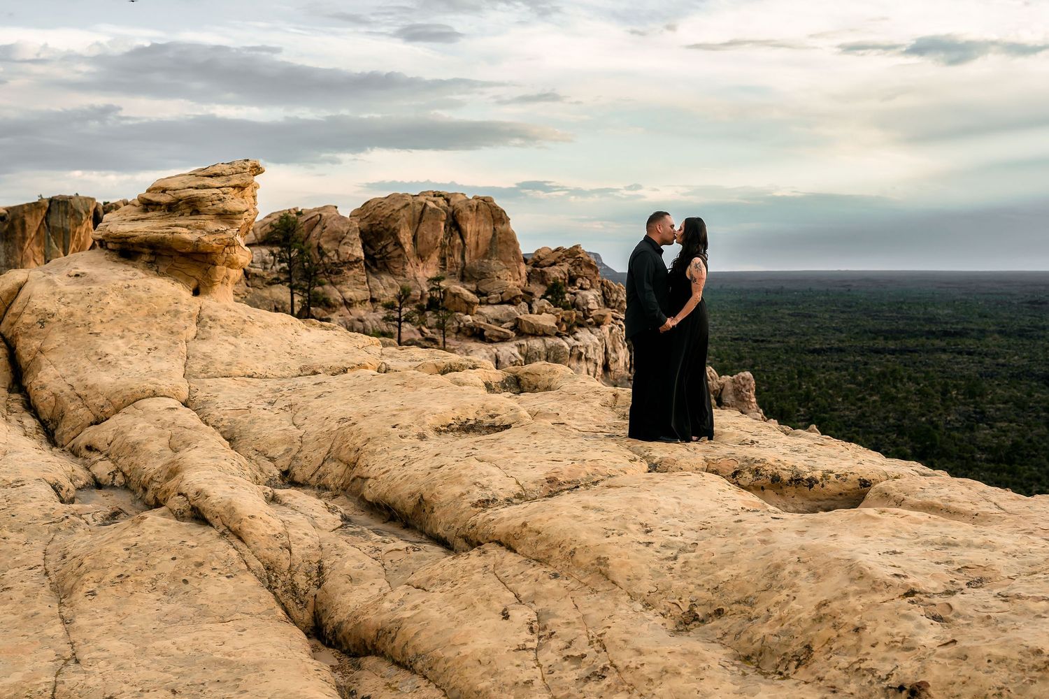Couple sharing a romantic kiss on rugged sandstone rock formation with sweeping desert landscape views.