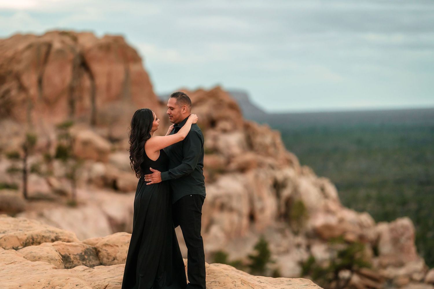 Couple embracing on rocky terrain with dramatic sandstone formations and forest backdrop.