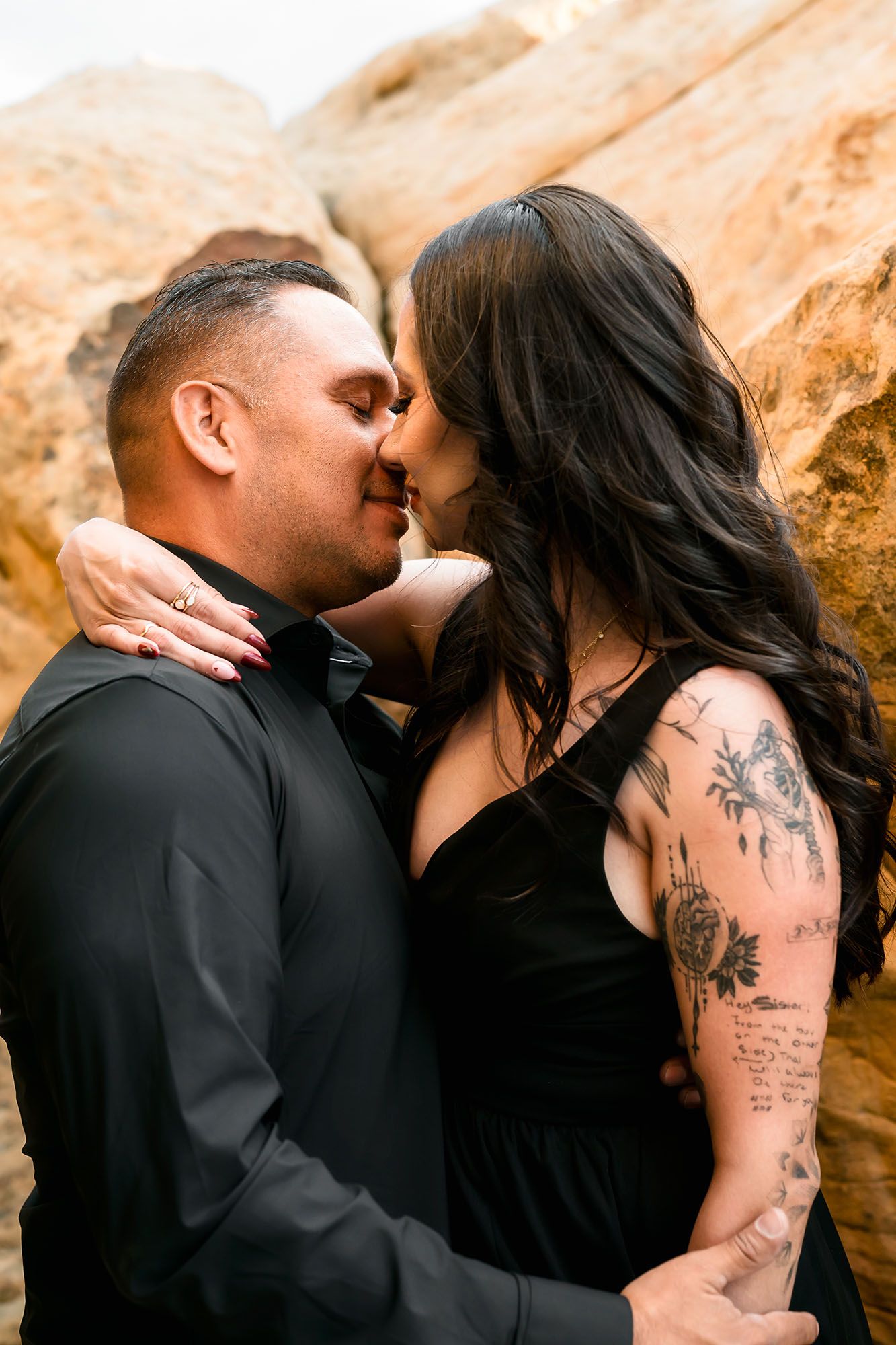 Couple kissing tenderly outdoors, woman with sleeve tattoos in black dress embracing man in black shirt, sandy rock backdrop.