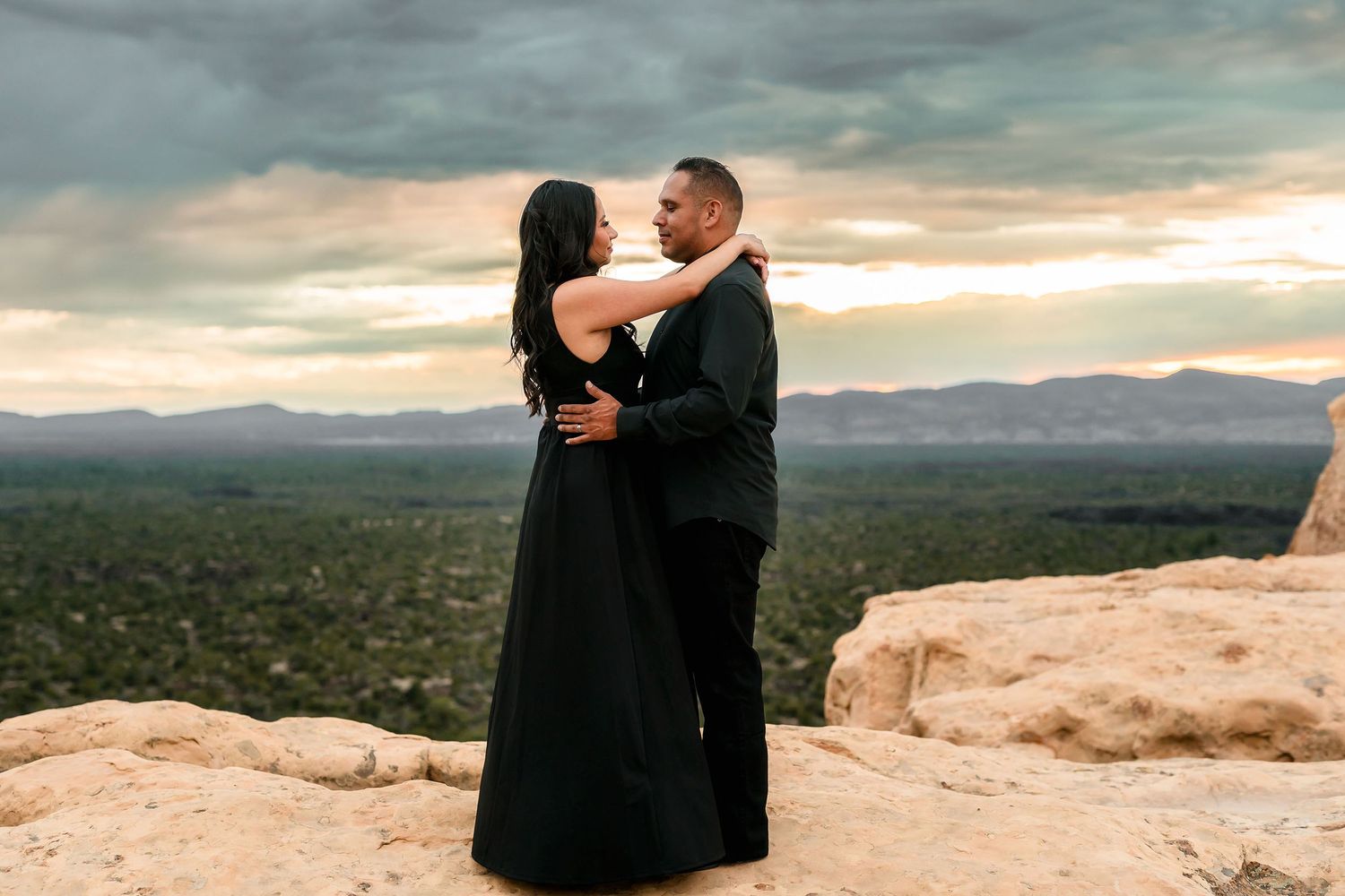 Romantic couple embracing in black attire on cliff edge with dramatic sunset sky.
