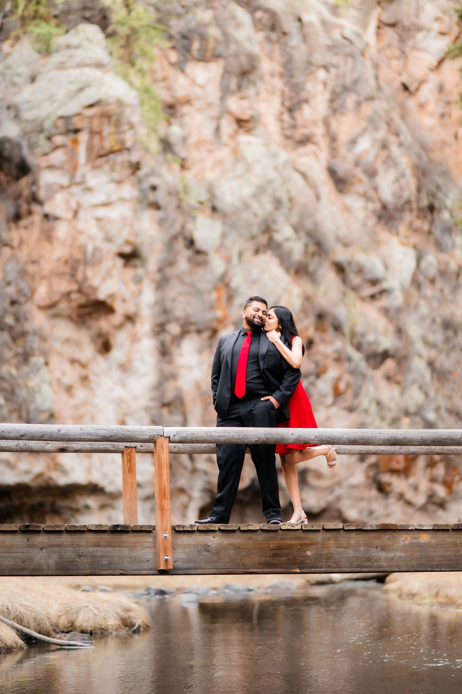 A romantic couple in formal attire shares an intimate moment on a wooden bridge with rocky cliffs in the background.