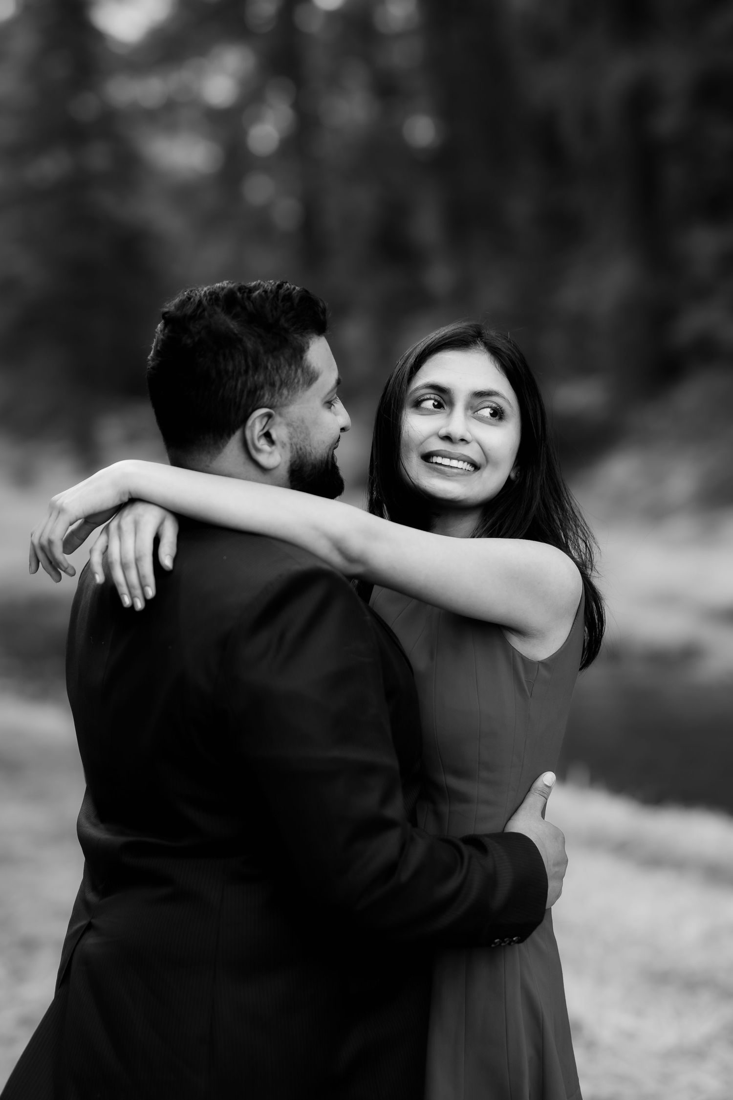 A couple shares a tender embrace outdoors in formal attire captured in black and white.