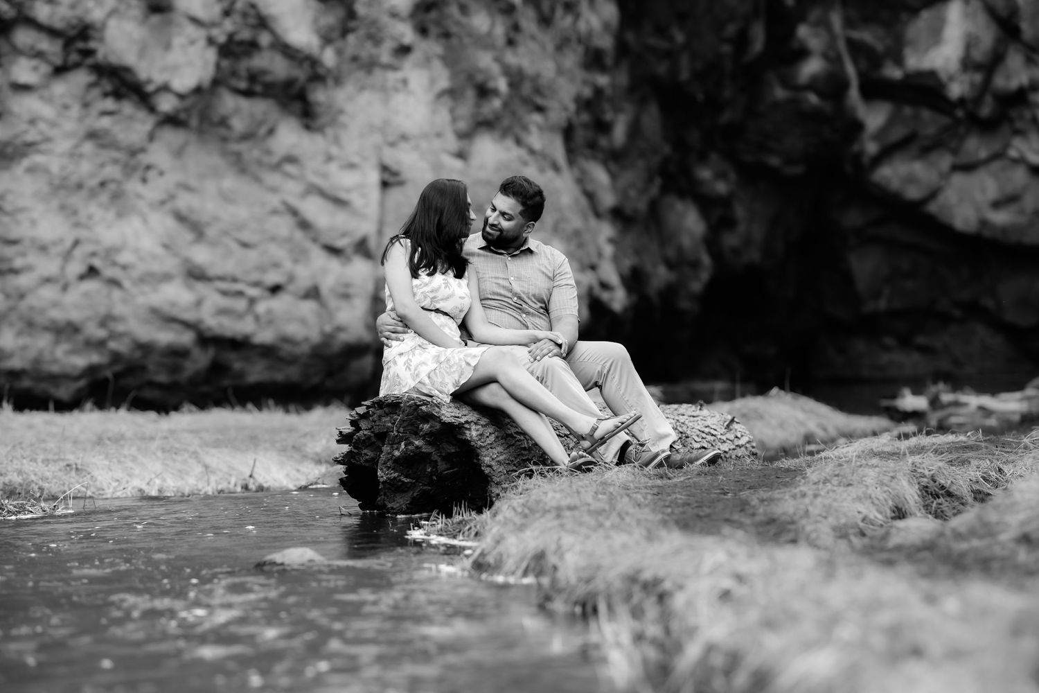 Black and white photo of couple sitting together on rocky outcrop by water.