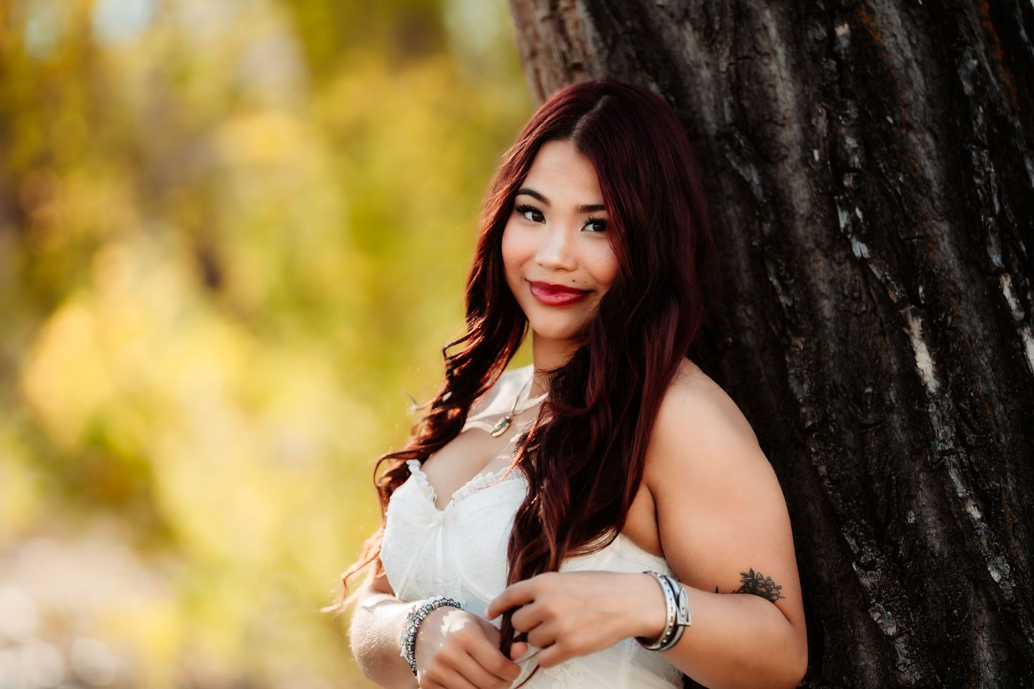 A woman in a white dress leans against a tree trunk with autumn foliage in the background.
