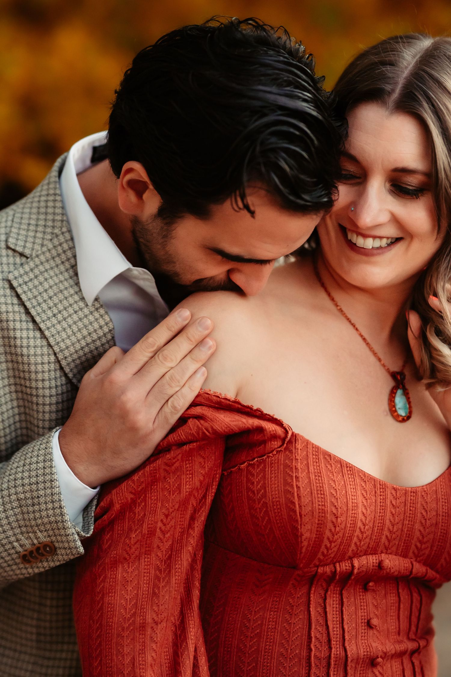 Romantic couple embraces tenderly, with person in red sweater and checkered shirt against fall foliage.