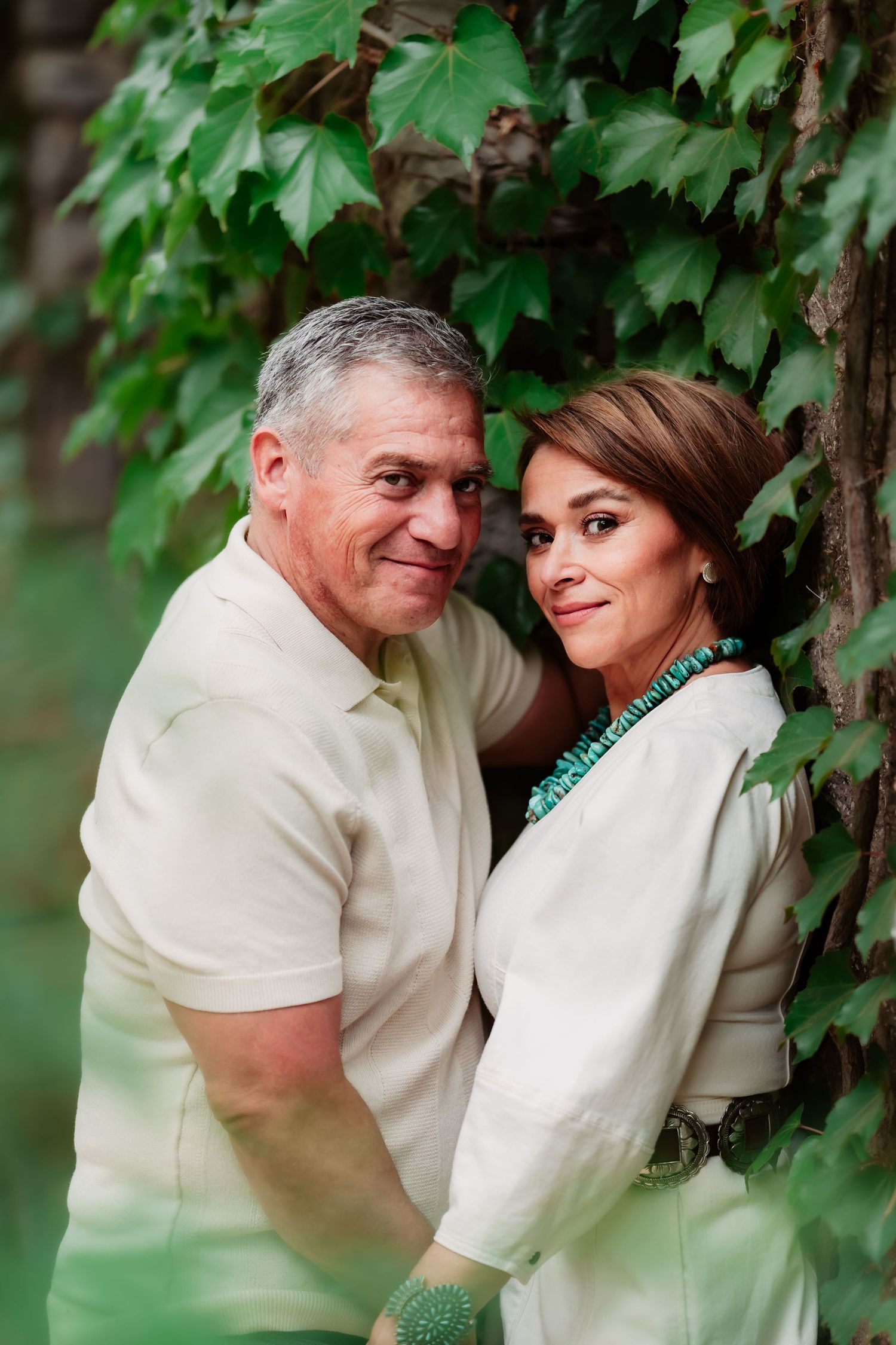 A couple in white outfits share intimate moments among green foliage in a romantic outdoor photoshoot.