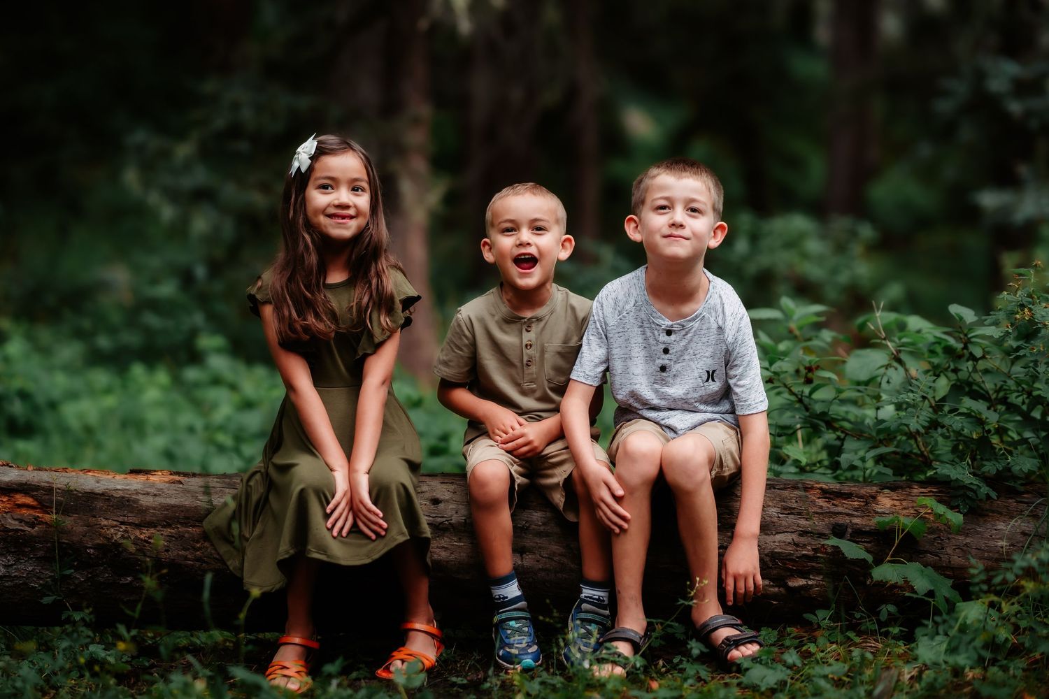 Three children sit together on a fallen log in a lush green forest setting.