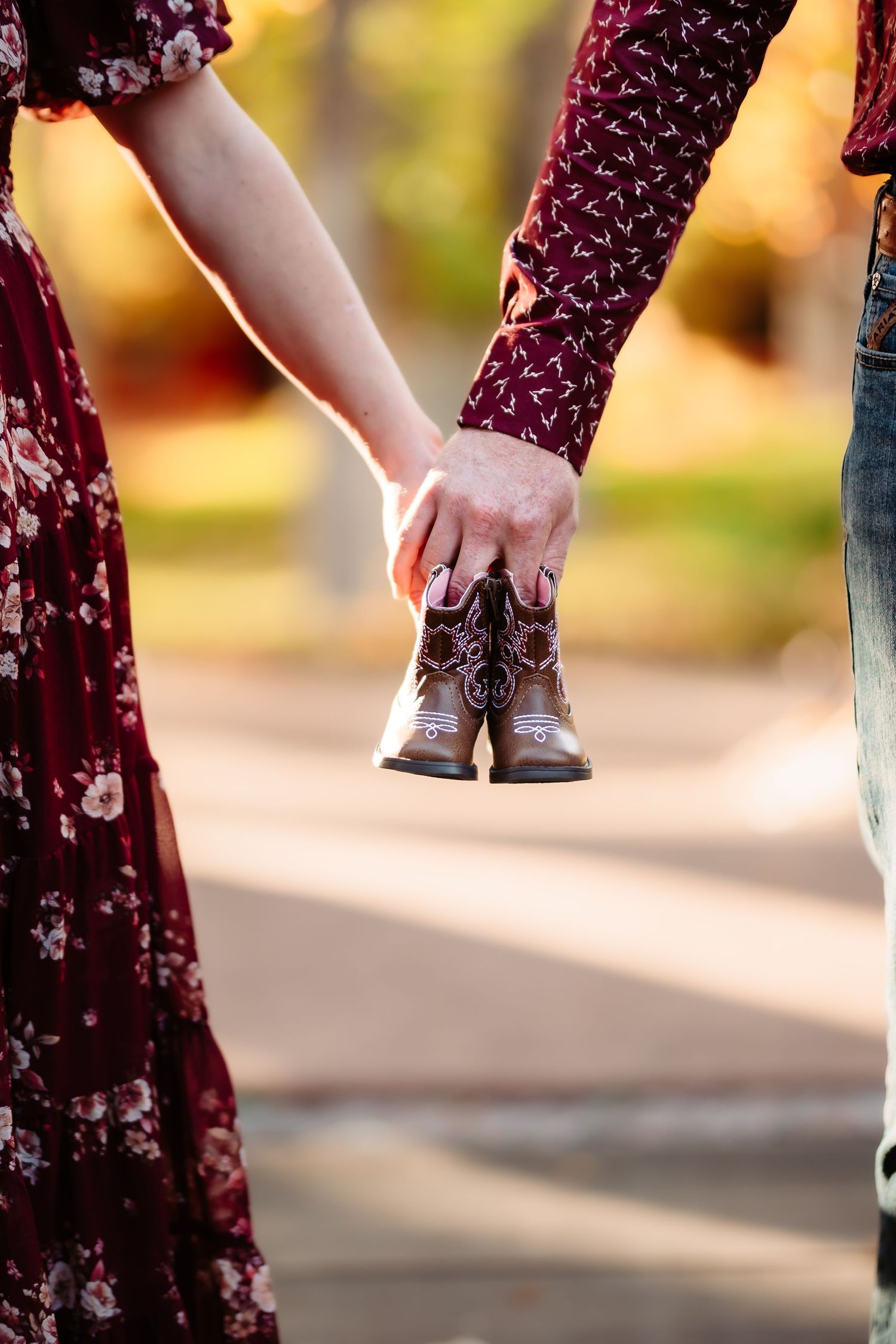 Woman in burgundy floral dress adjusts brown leather boots while standing outdoors in autumn sunlight.