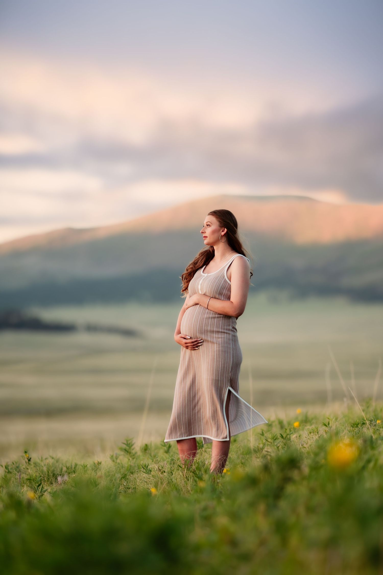 A pregnant woman in a flowing white dress stands in a green meadow at sunset with mountains in the background.