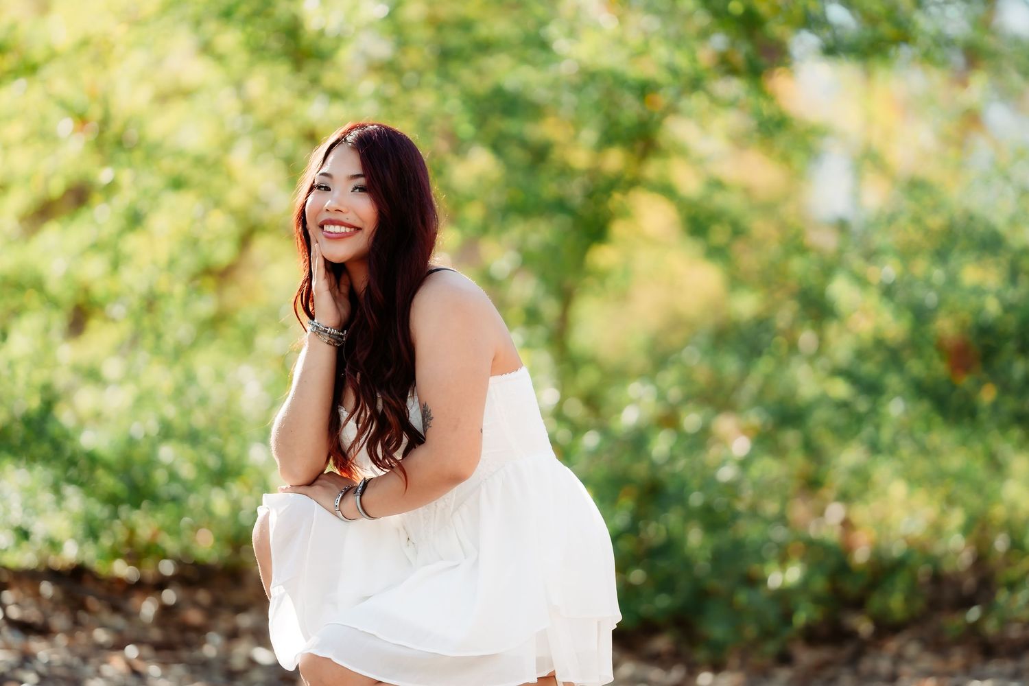 A person in a white dress poses outdoors with natural greenery and trees in the background.
