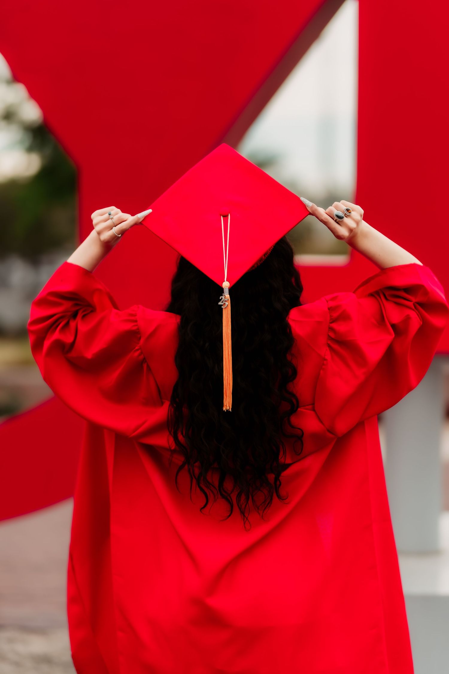 Graduate in bright red cap and gown shows back view while holding graduation cap against red background.