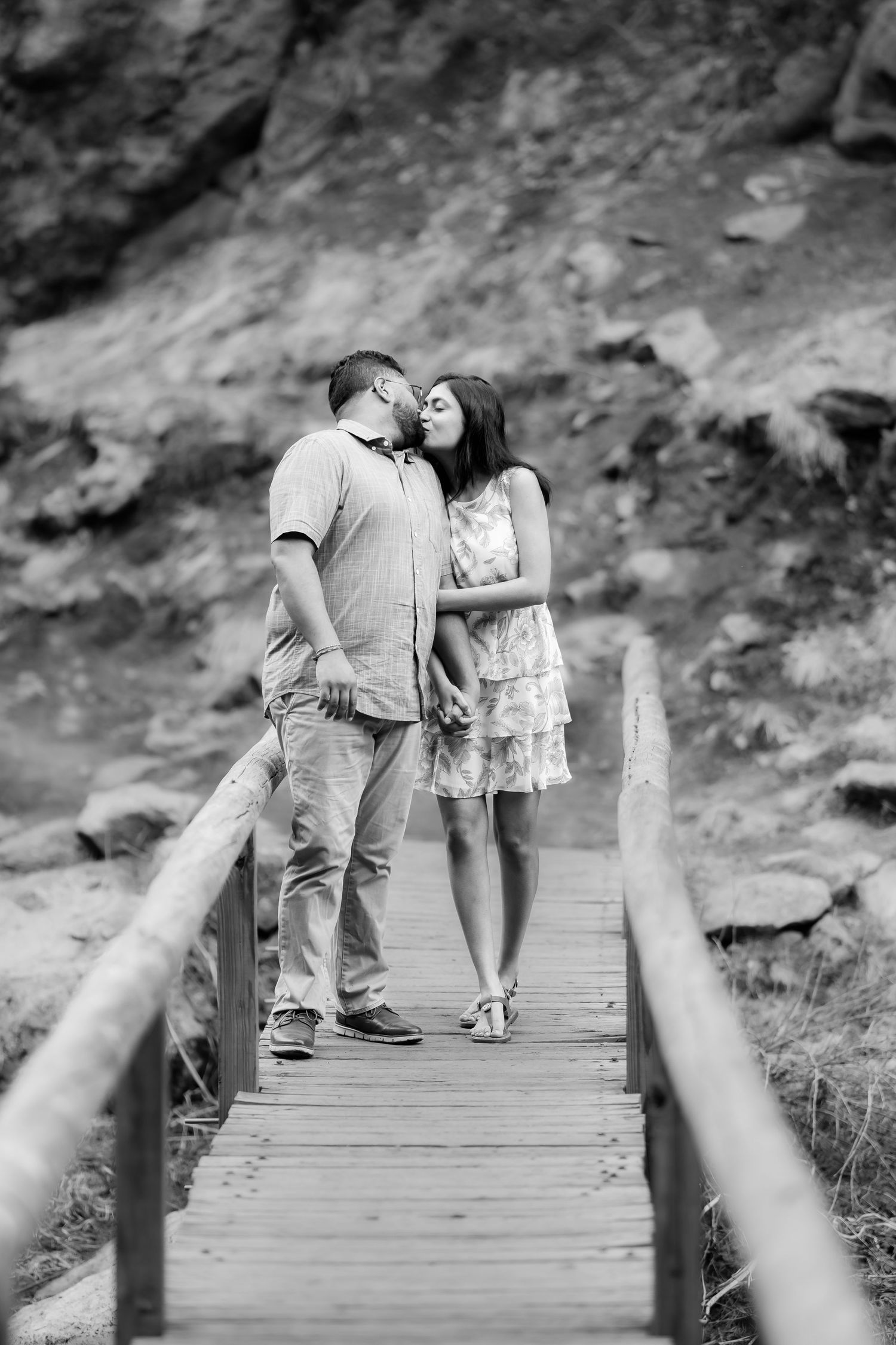 A romantic couple embrace while standing on a wooden boardwalk against a rocky backdrop in black and white.
