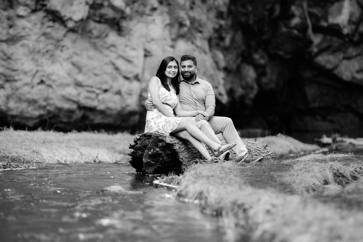 Black and white photo of couple sitting together by a stream against rocky backdrop.