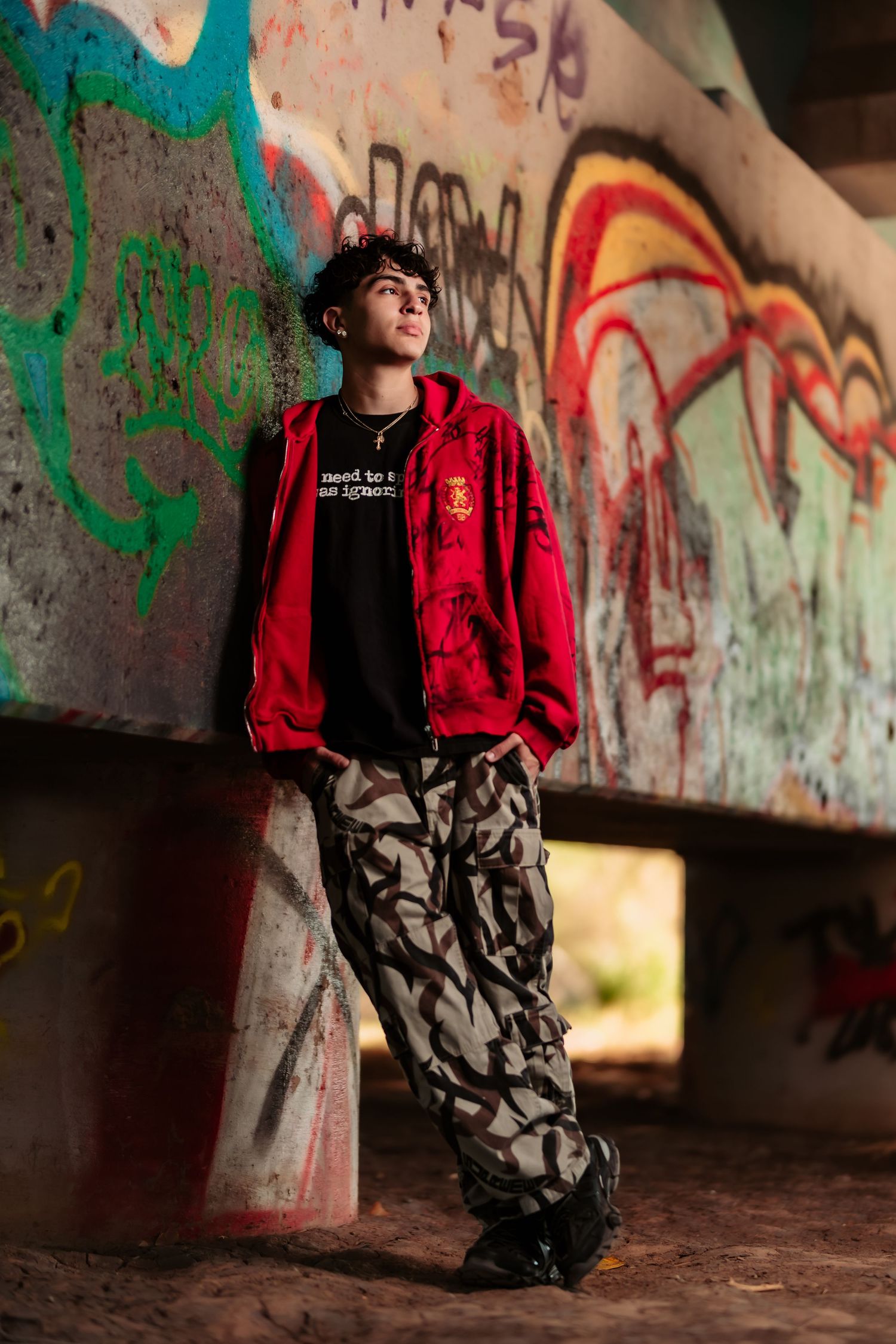 Urban fashion portrait of someone in a red jacket and camo pants posing against graffiti-covered wall.