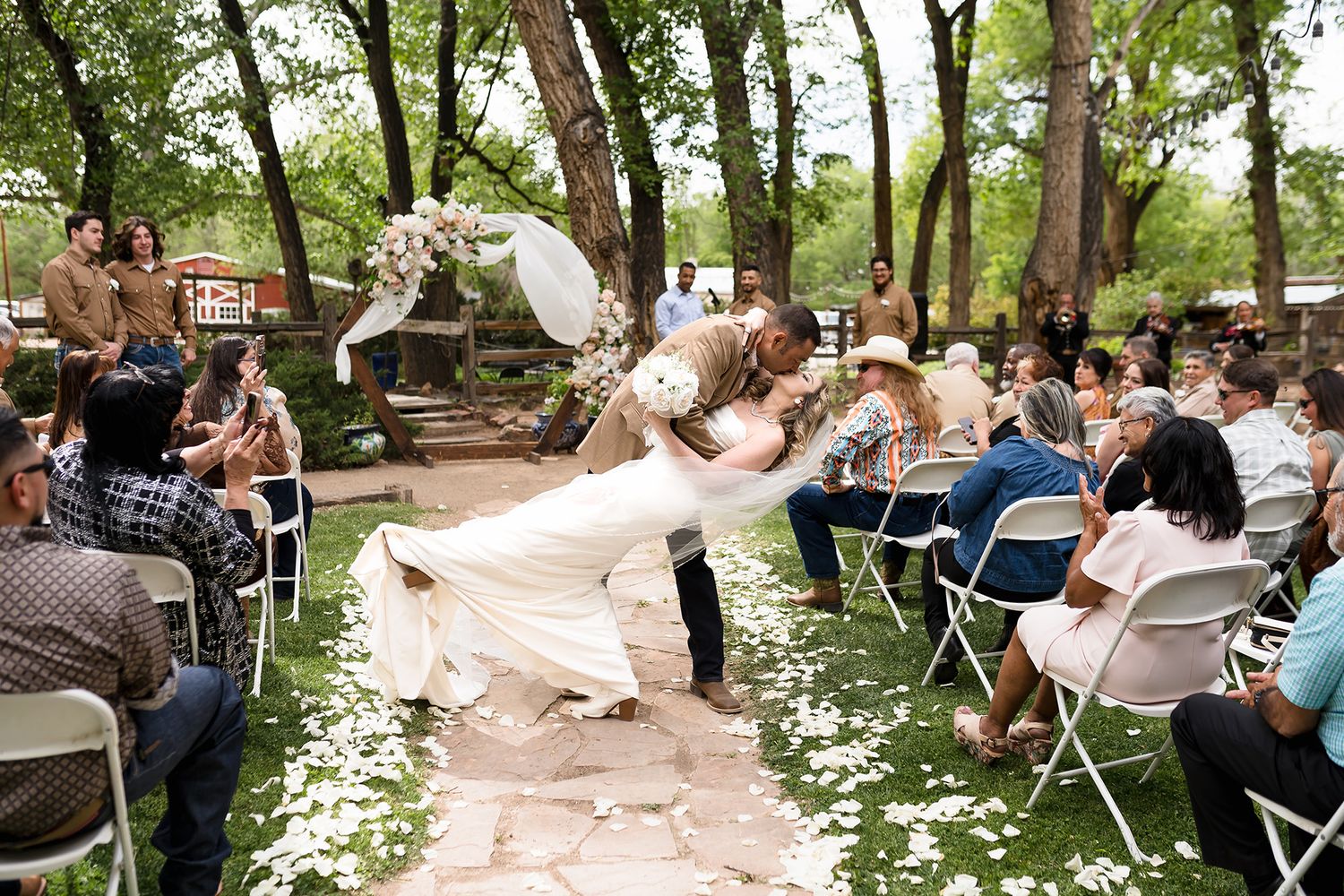 Groom dips bride in white gown during outdoor wedding ceremony with guests seated on white chairs amid lush green trees.