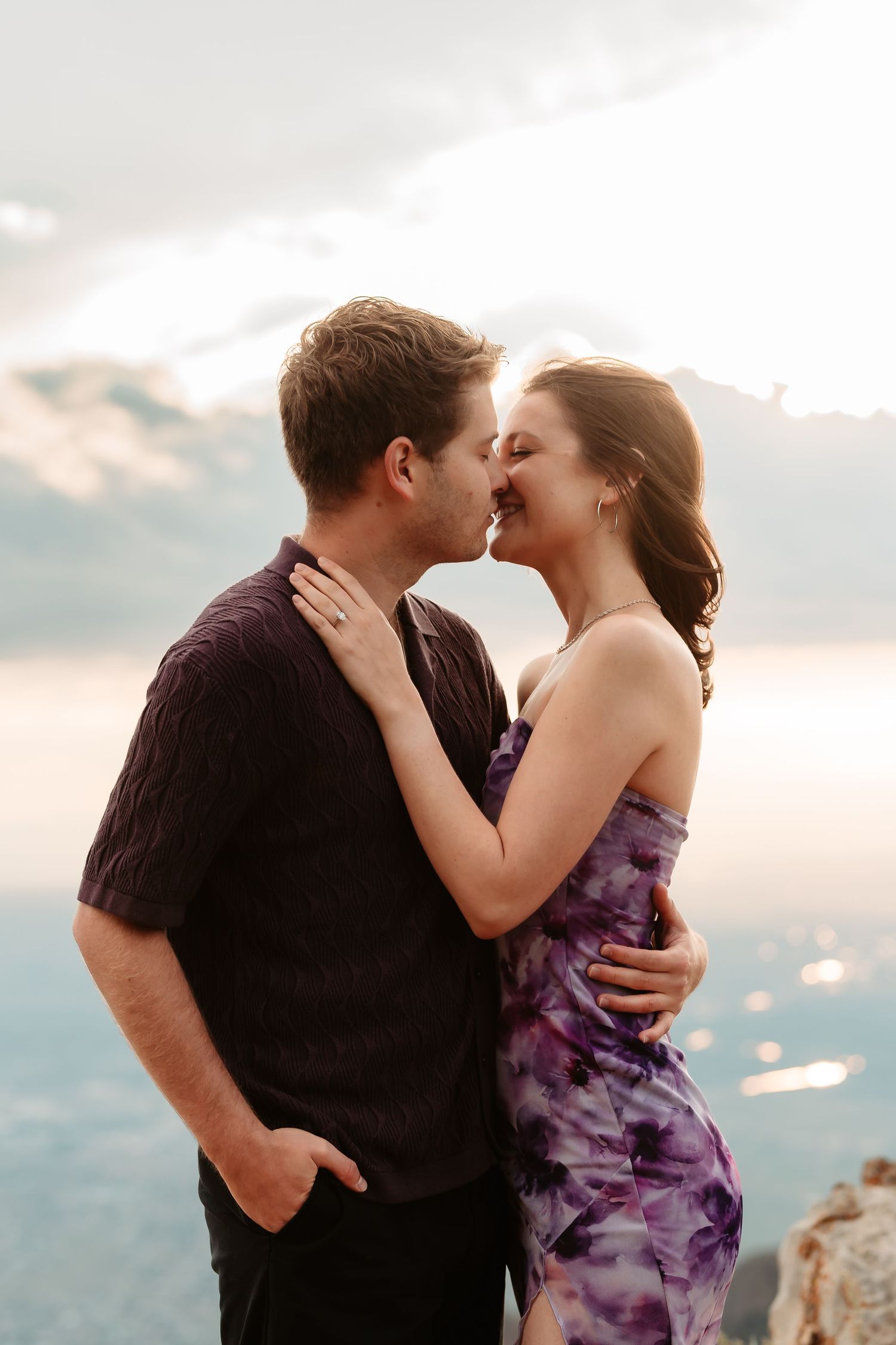 Two people embracing and sharing a romantic kiss on a mountain cliff at sunset.