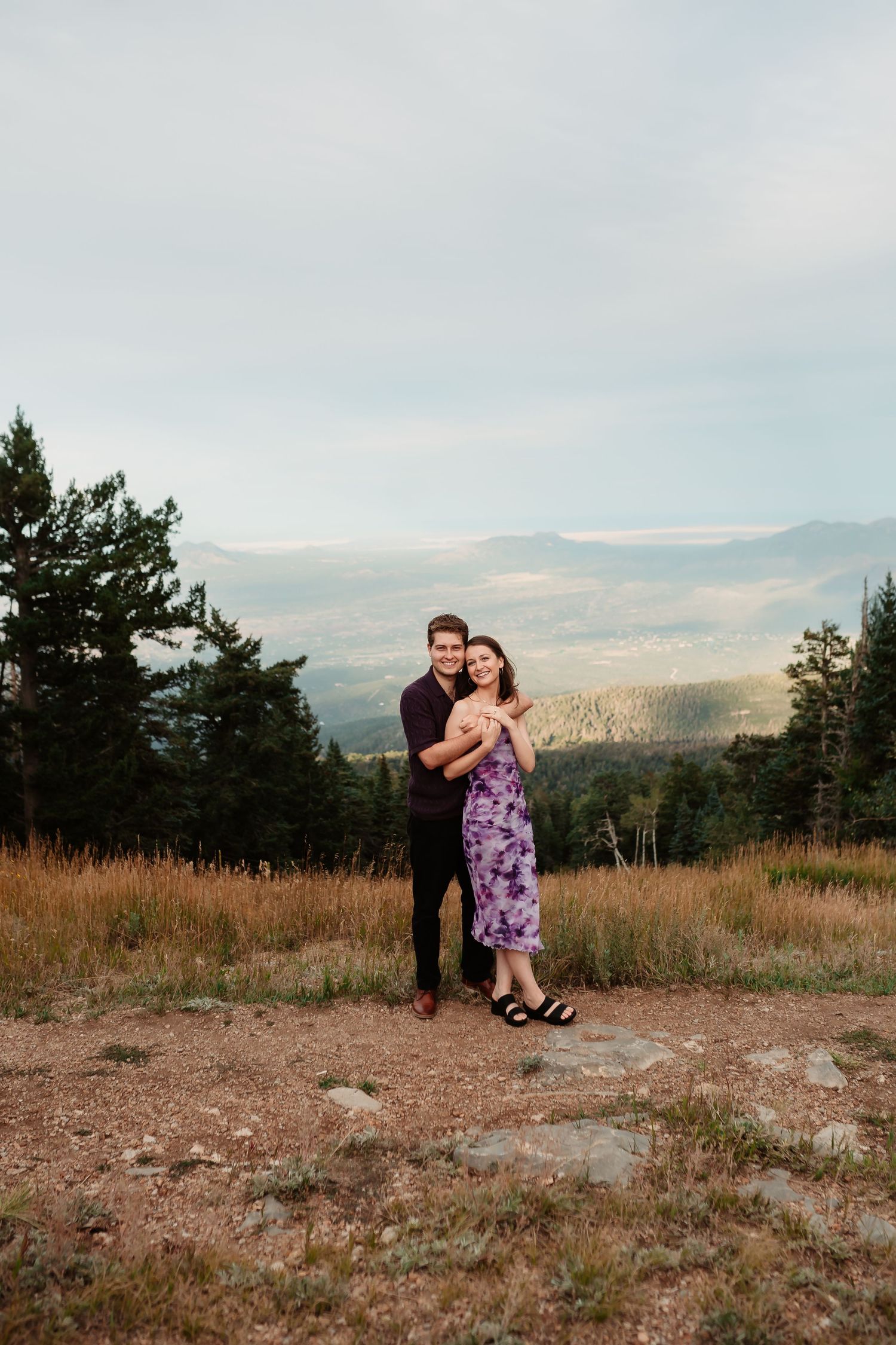 A couple shares an intimate moment on a mountaintop with a scenic valley vista behind them.