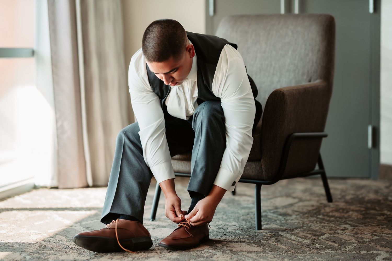 A person in brown leather shoes sits in an armchair while tying their laces in a minimal indoor setting.