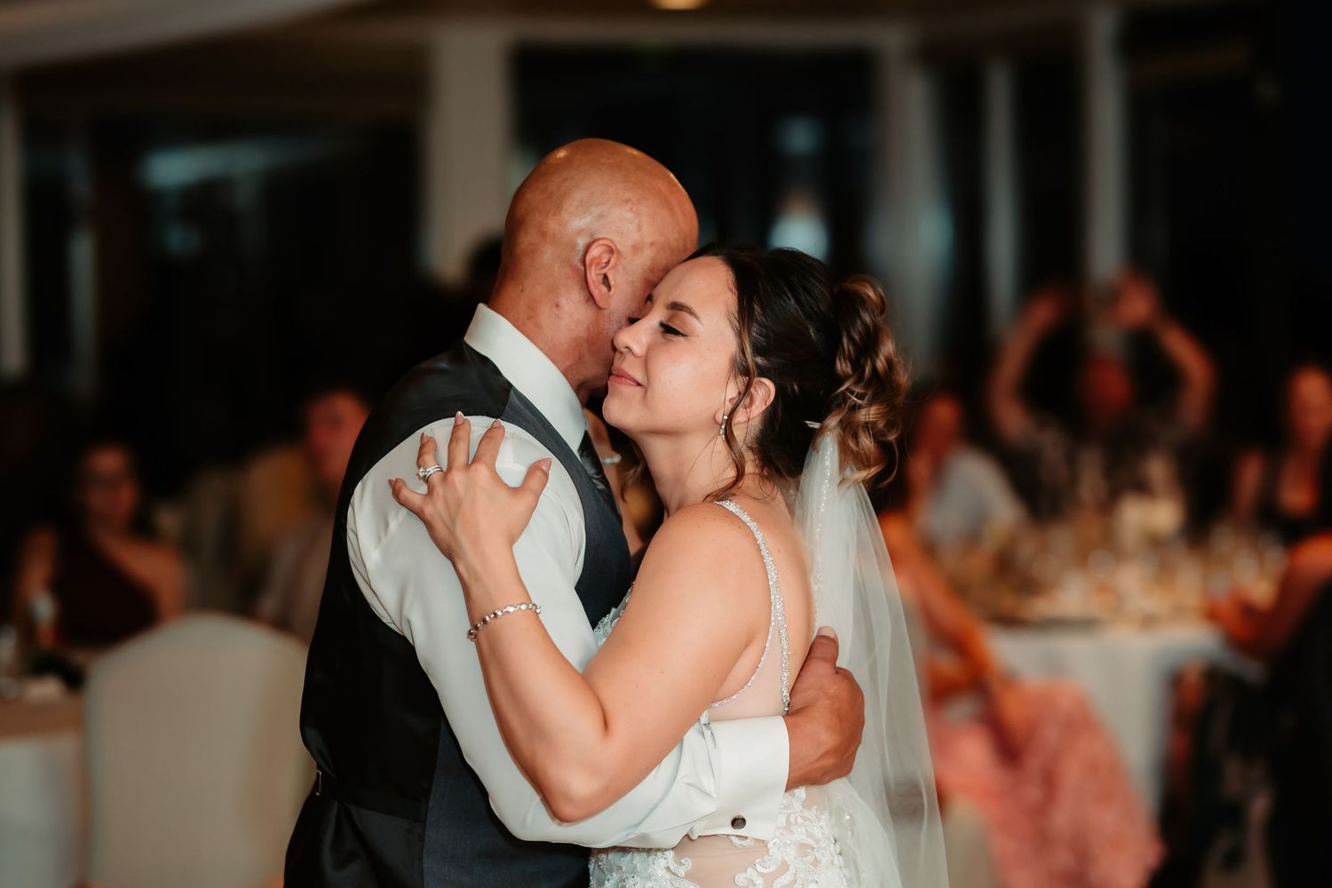 Newlyweds share a romantic slow dance moment while reception guests watch in the background.