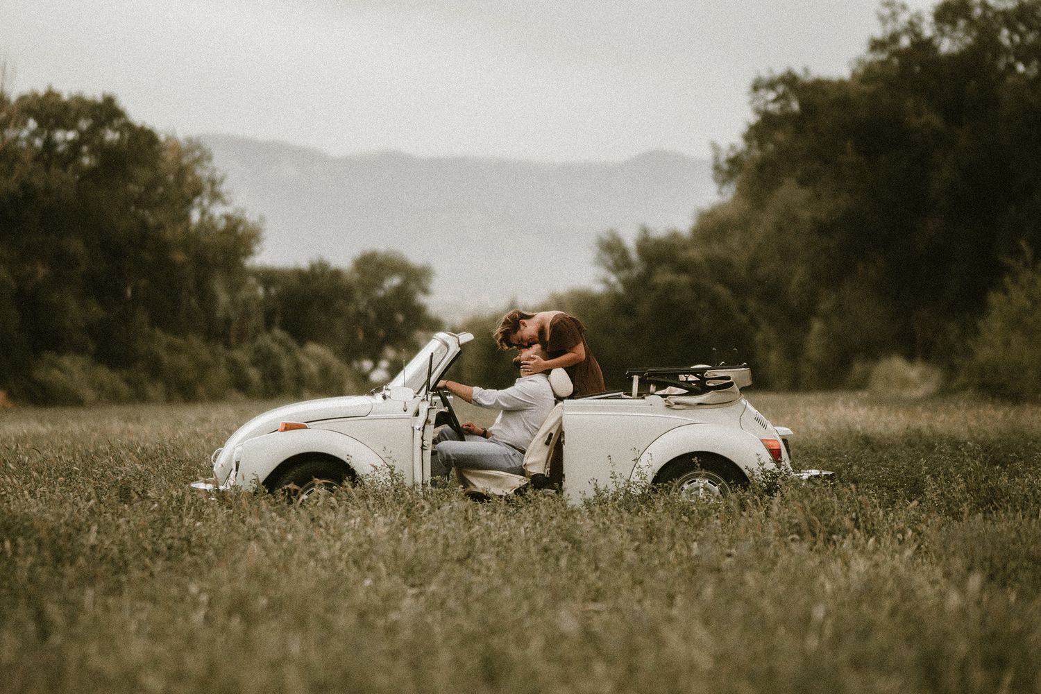 Couple kissing in a white vintage VW Beetle convertible parked in a green meadow with mountains in the background.