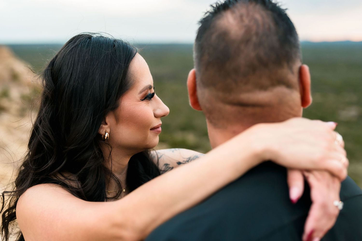A woman with dark hair embraces a man outdoors, gazing at him with rocky desert landscape behind them.