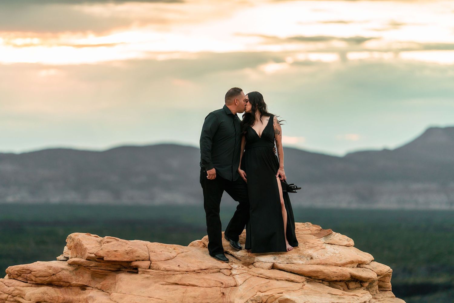 Couple sharing a kiss on a sandstone rock at sunset with mountain range behind them.