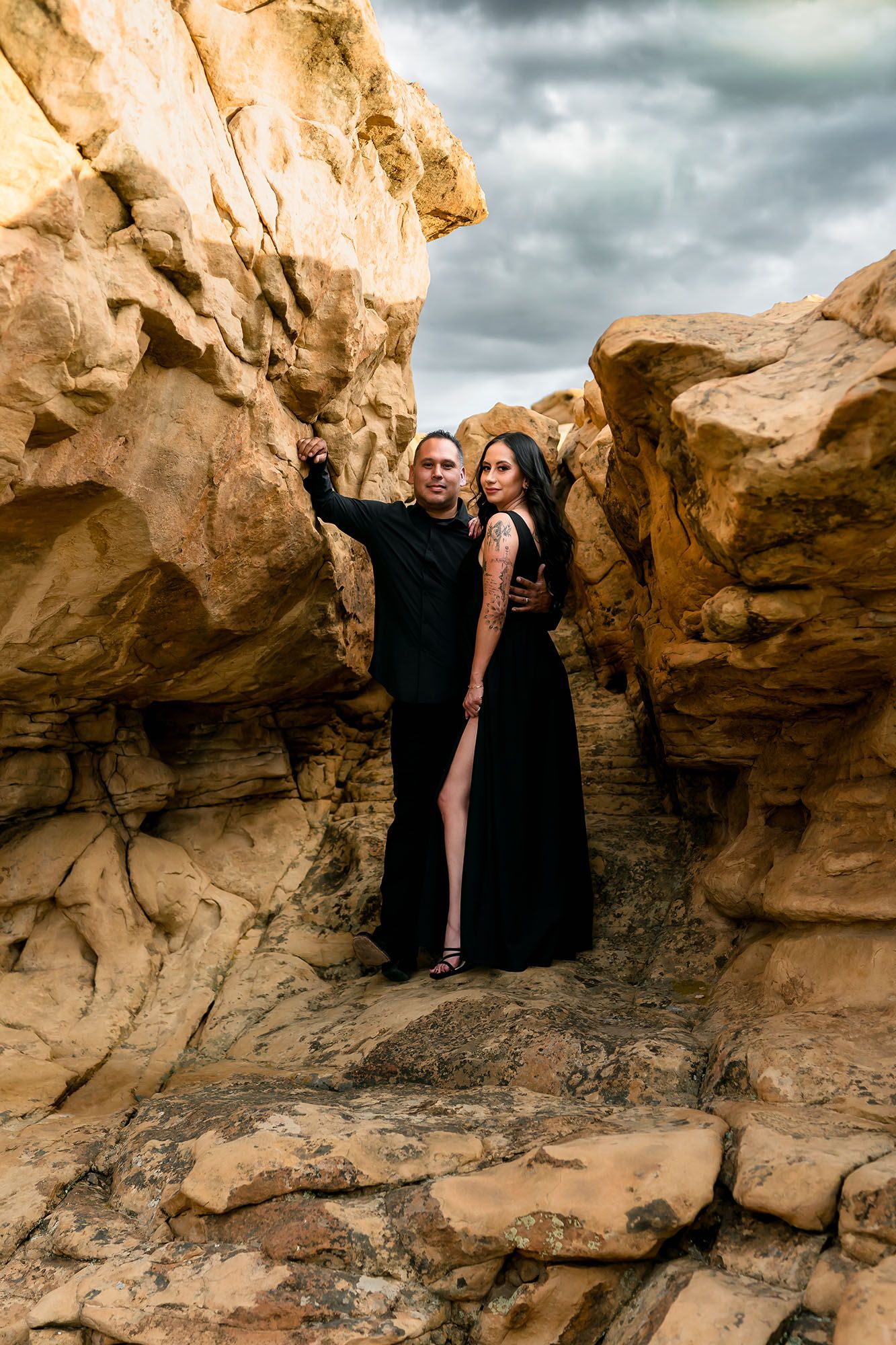 Couple in black formal wear posing between golden sandstone rock formations under dramatic cloudy sky.