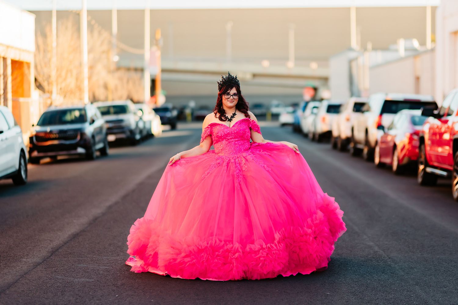 A bright pink quinceañera dress stands out against an urban backdrop with parked cars and buildings.