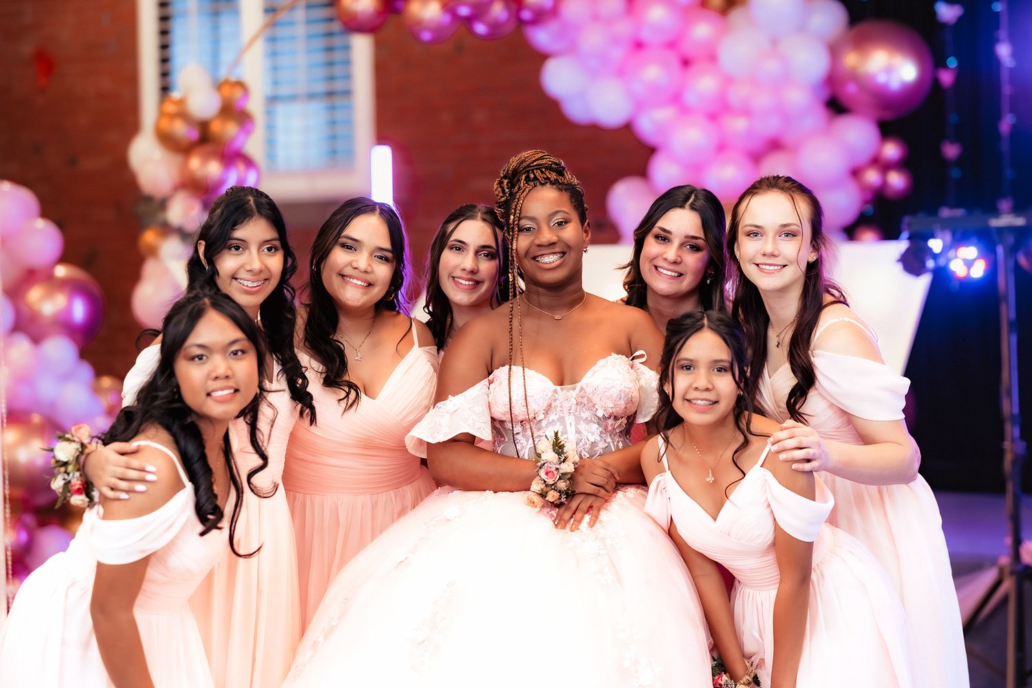 Group wearing white dresses pose together at formal event with pink and blue lighting in background.
