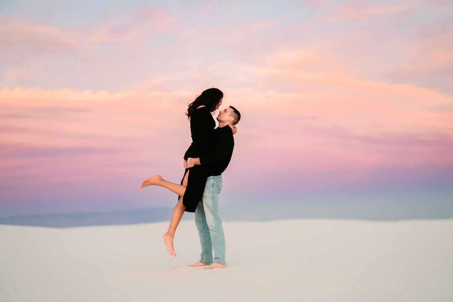 A couple silhouetted against a pink and purple sunset sky performs a romantic dance lift pose on the beach.