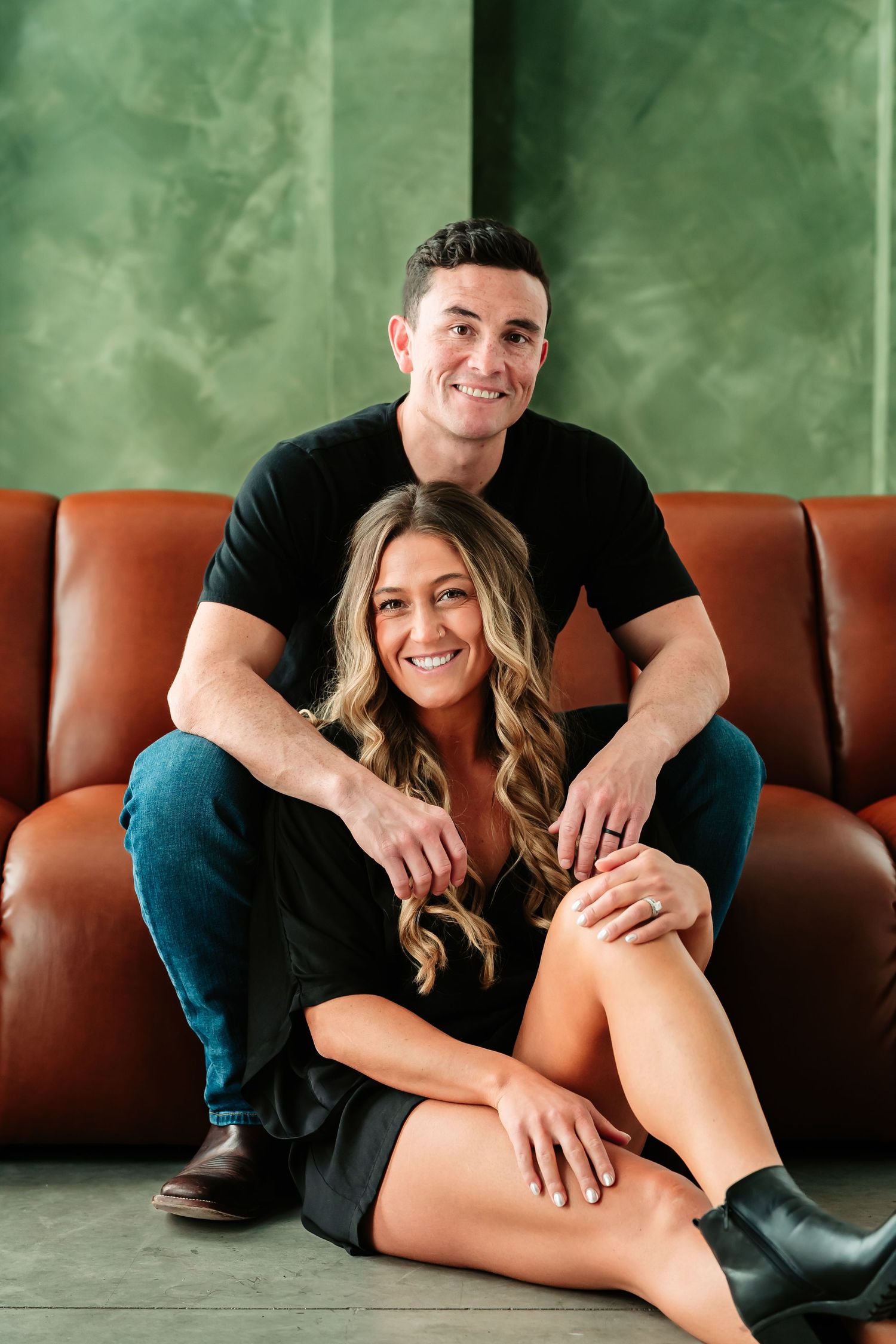 A couple lounges on a brown leather couch in a studio setting with green textured walls.