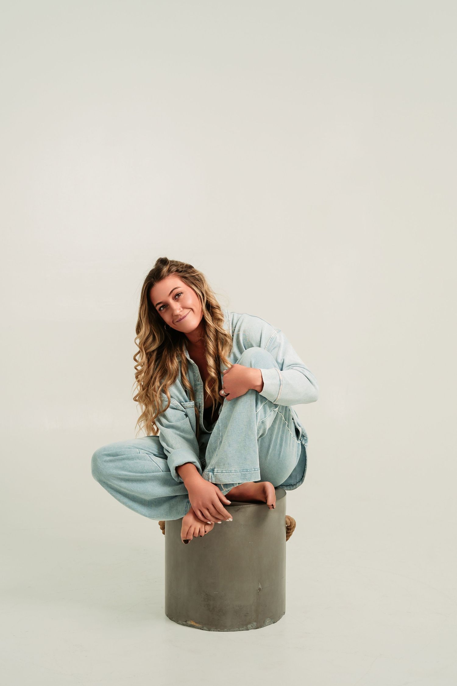 A person in a light blue top poses playfully while sitting on a gray cylindrical pedestal against a white background.