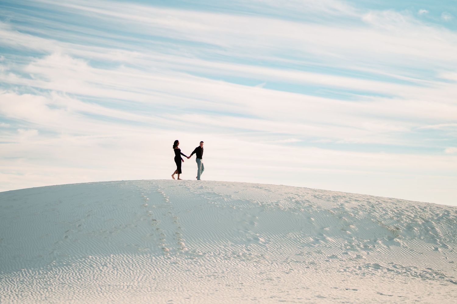 Two silhouetted figures stand together atop a white sand dune against a bright blue sky.